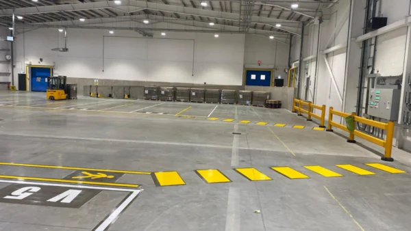 Yellow hatched pedestrian crossing and bay numbering marked on a depot floor beside forklift and safety barriers